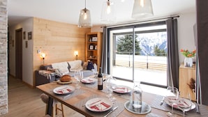 Un coin repas avec une table en bois dressée pour le repas, un canapé en arrière-plan et une vue sur les montagnes par une grande fenêtre.