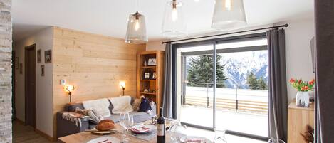 A dining area with a wooden table set for a meal, a sofa in the background, and a view of mountains through a large window.