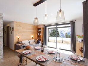 Ein Essbereich mit Holztisch, gedeckt für eine Mahlzeit, einer Couch im Hintergrund und Blick auf Berge durch ein großes Fenster.