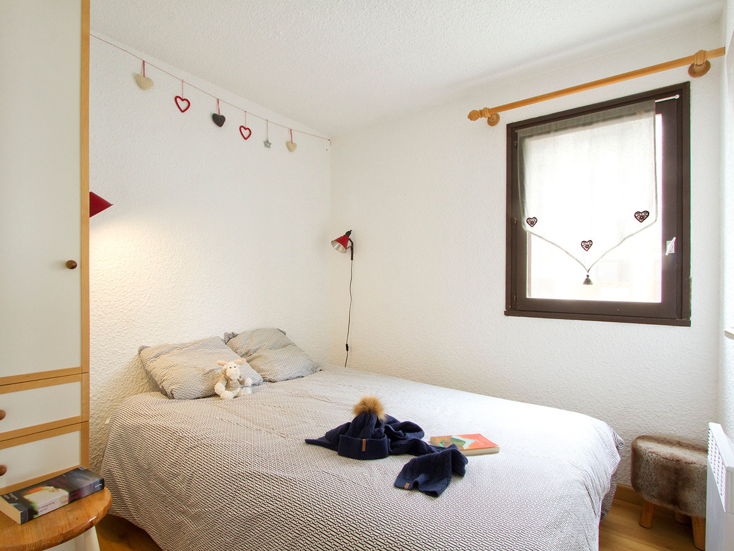 Un dormitorio con cama, ventana, lámpara y un taburete pequeño.