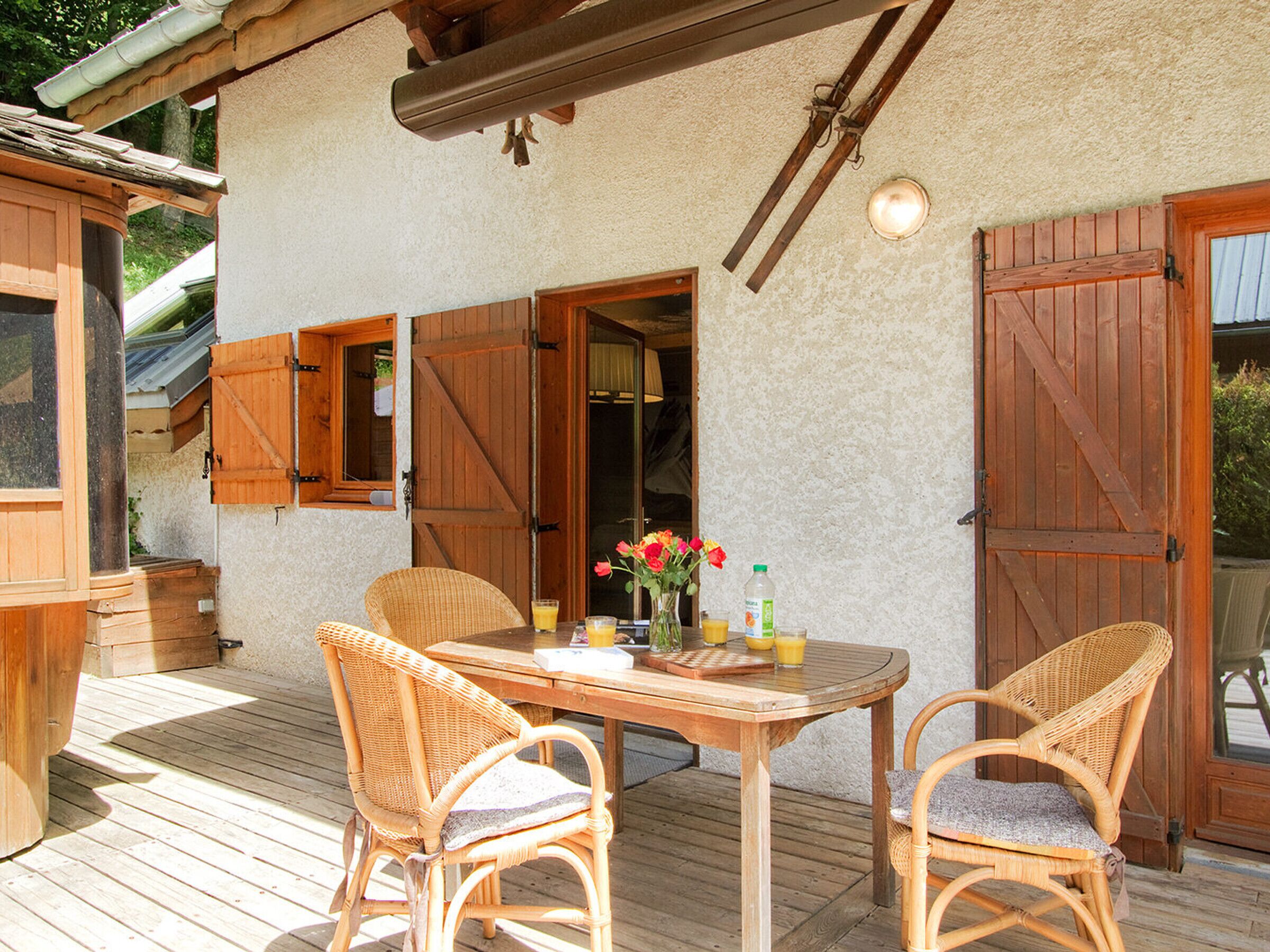 Una terraza de madera con una mesa y sillas de mimbre, una puerta de madera y un ventanal con contraventanas.