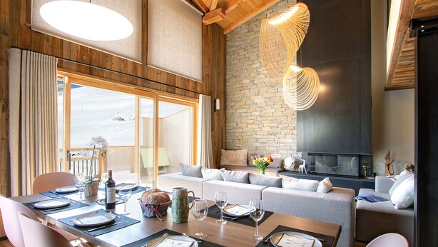 Un comedor con una mesa de madera dispuesta para una comida, una chimenea de piedra y un amplio ventanal con cortinas.