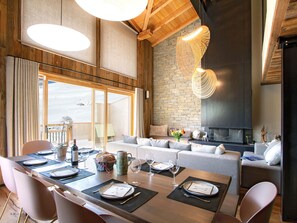 Un coin repas avec une table en bois dressée pour le repas, une cheminée en pierre et une grande fenêtre avec des rideaux.
