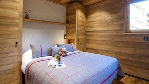 Interior de una cabaña de madera con una cama, almohadas, un osito de peluche y una ventana.