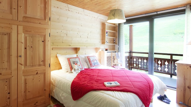 Ein Schlafzimmer mit Holzwandverkleidung, einem Bett, Kissen, einer roten Decke, einem Nachttisch und einem Fenster mit Blick ins Freie.