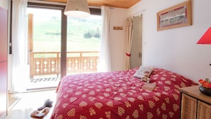 Ein Schlafzimmer mit einem Bett, Holzboden, einem fenster mit Vorhängen, einer Holztür und Blick ins Freie.