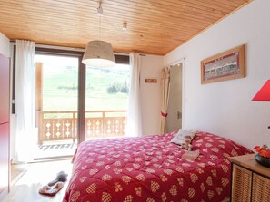 Ein Schlafzimmer mit einem Bett, Holzboden, einem fenster mit Vorhängen, einer Holztür und Blick ins Freie.