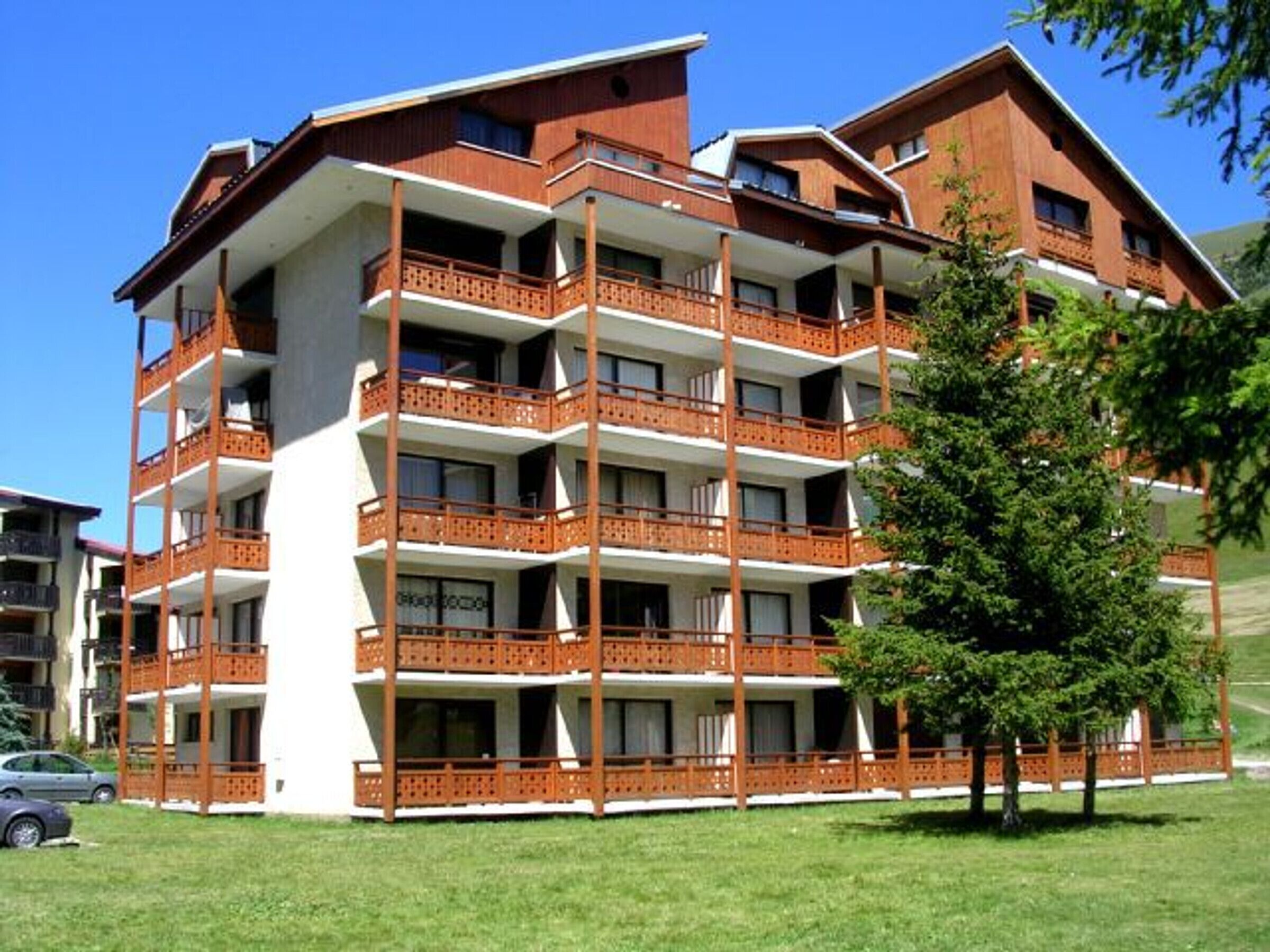 Un hotel de varios pisos con balcones, un coche estacionado al frente y un árbol cercano.
