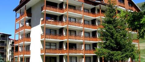 Un hotel de varios pisos con balcones, un coche estacionado al frente y un árbol cercano.
