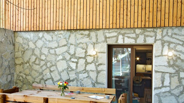Una mesa de madera con bancos y un jarrón de flores frente a una pared de piedra con un ventanal que da a una zona de cocina.