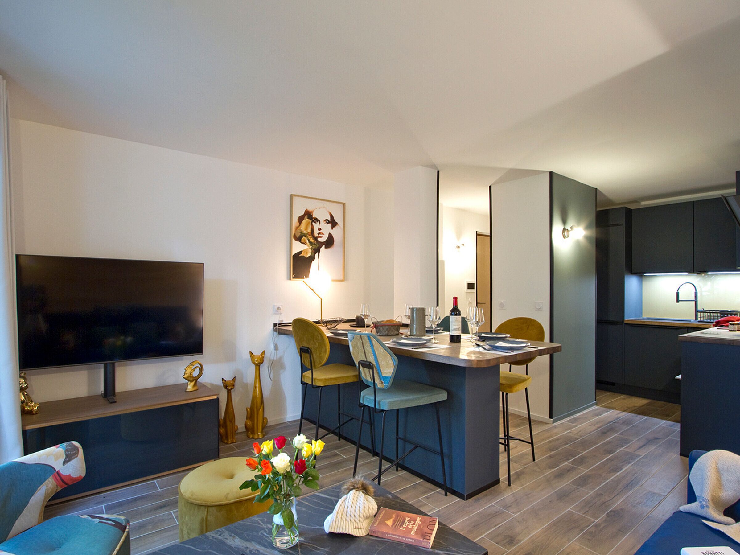 Ein modernes Wohnzimmer mit Flachbildfernseher, eine Essecke mit Barhocker und eine Küche mit Einbauherd.