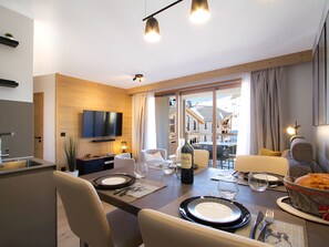 A modern dining area with a wooden table set for two, a television mounted on the wall, and a view of the outdoors through large windows. - Apartment with slope view - 5 stars (Huez)