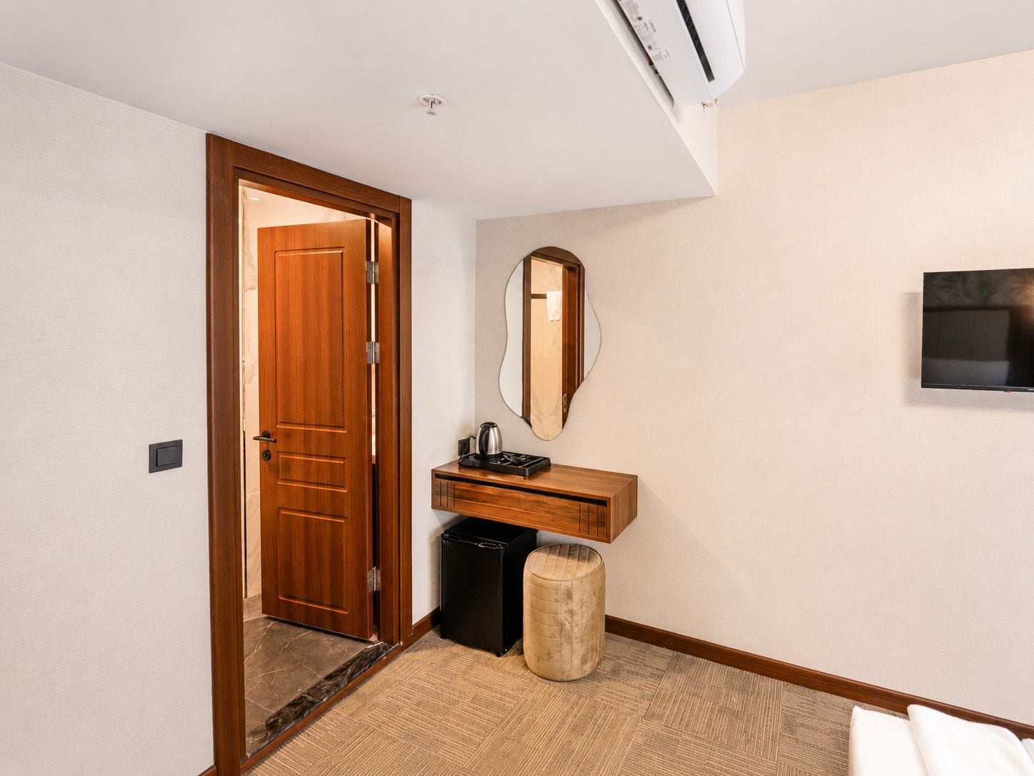 Ein Hotelzimmer mit Holztür, einem kleinen Schminktisch mit Spiegel, einer schwarzen Kaffeemaschine, einem schwarzen Mülleimer und einem runden Hocker.