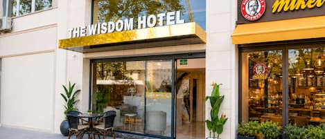 A hotel exterior with a sign reading 'The Wisdom Hotel' and a visible coffee shop named 'Mikel' inside.