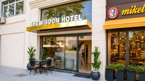 Exterior de un hotel con un cartel que dice 'The Wisdom Hotel' y una cafetería visible llamada 'Mikel' en su interior.