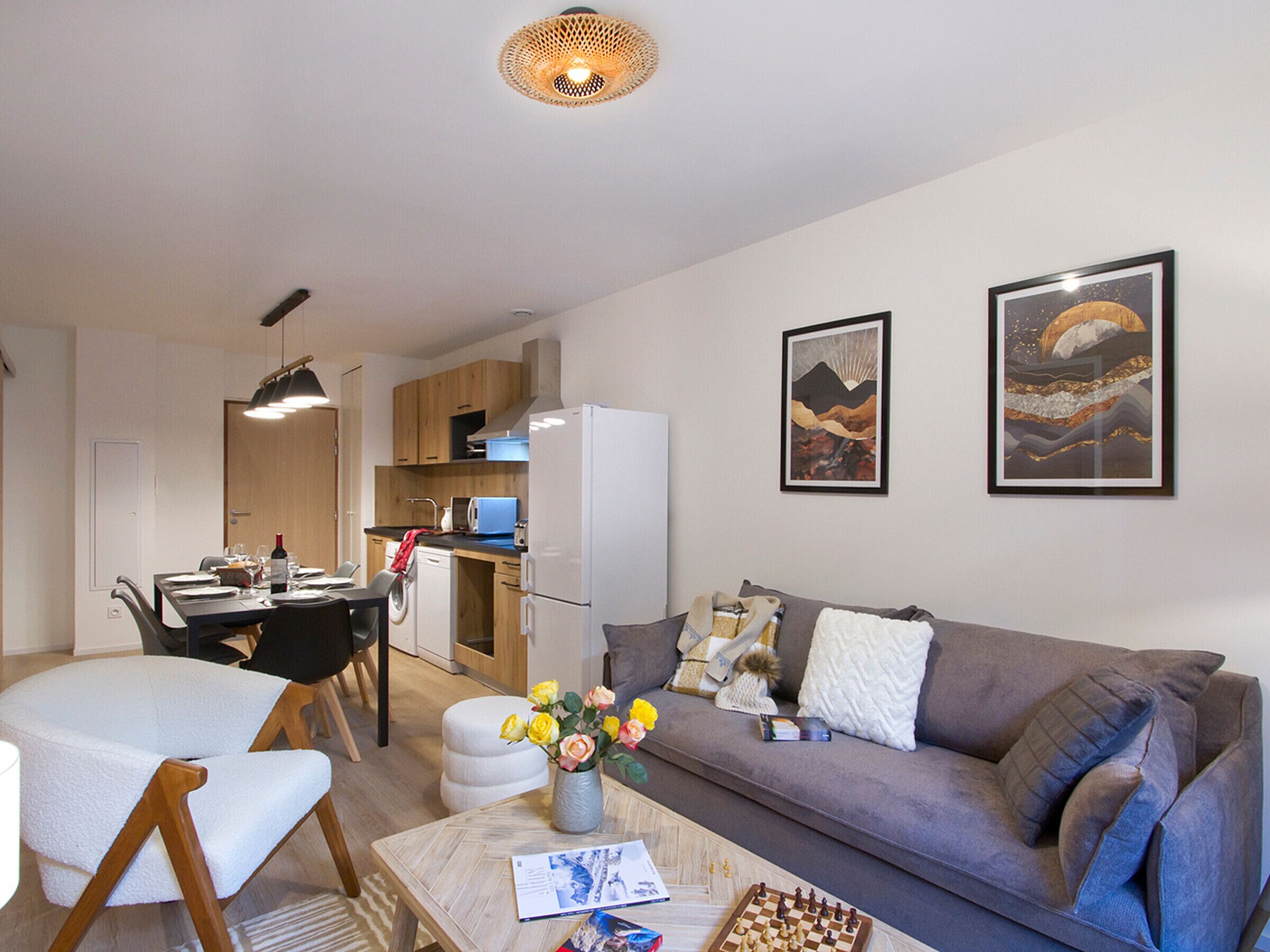 A modern living room with a sofa, coffee table, and a chessboard. There is a dining area with a table and chairs, and a kitchen with cabinets and appliances.