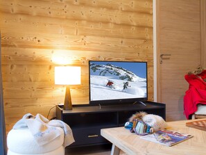 A wooden-paneled room with a television displaying a snowy mountain scene, a lamp, a table with a magazine, and a red jacket on a chair. - Beautifully decorated apartment, calm - 4 guests (Les Deux Alpes)