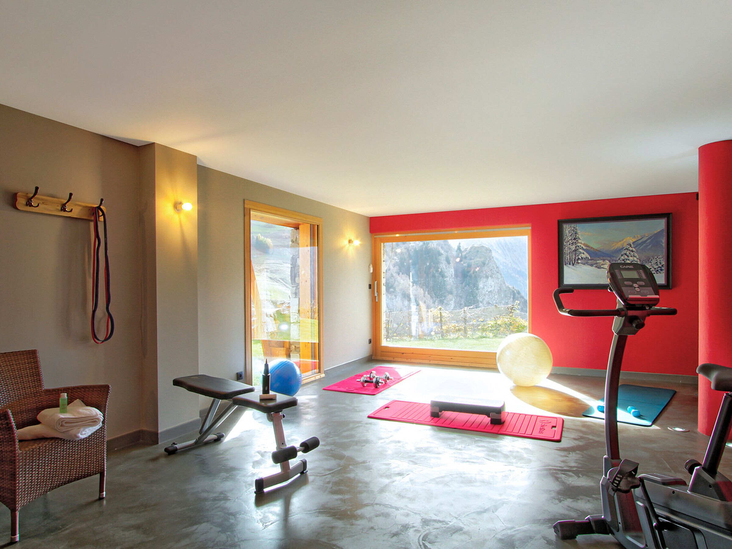 Ein Fitnessstudio mit Trainingsgeräten, einer Bank, einem Rudergerät und einem großen Fenster mit Blick auf die Berge.