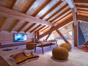 Ein gemütliches Dachzimmer mit Holzbalkendecke, einem Fernseher, einem Tisch und zwei gestrickten Hocker.