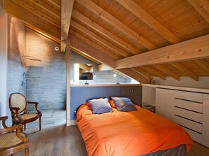 Ein Schlafzimmer mit geneigter Decke, einem Bett mit orangefarbener Bettwäsche, einem Holzstuhl und einem kleinen Fenster.