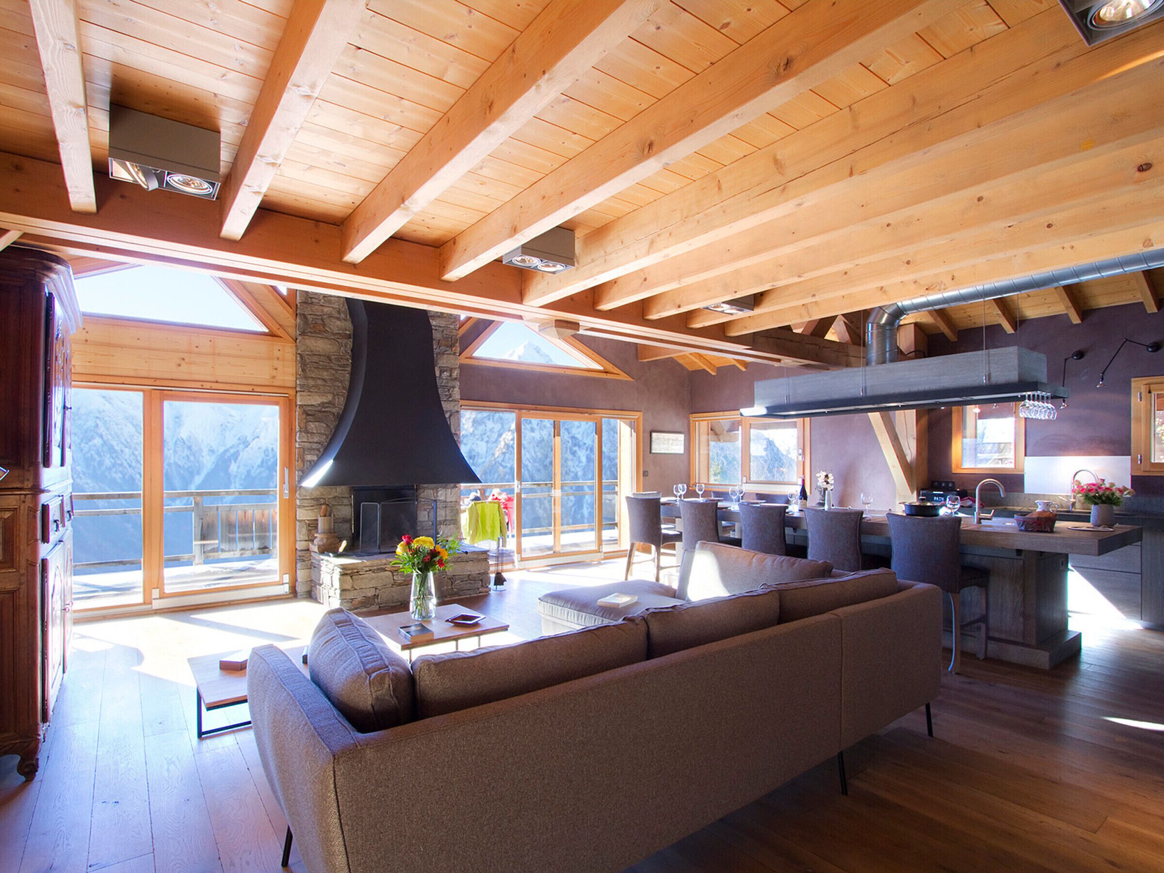 A spacious living area with a large wooden ceiling, a stone fireplace, and a view of the mountains through large windows.