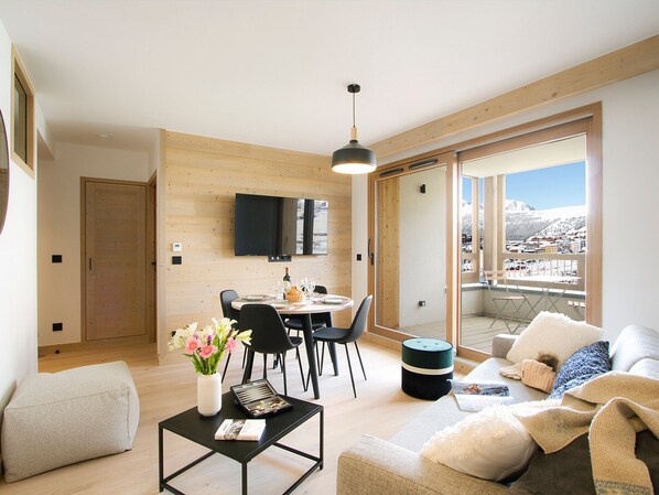 En moderne stue med spiseplads, et fladskærms-TV monteret på væggen og en balkon med udsigt over et sneklædt landskab.