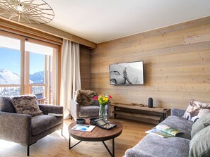 A modern living room with a large window, wooden walls, and a flat-screen TV mounted on the wall. - Spacious apartment ski-in/ski-out - 8 guests (Huez)