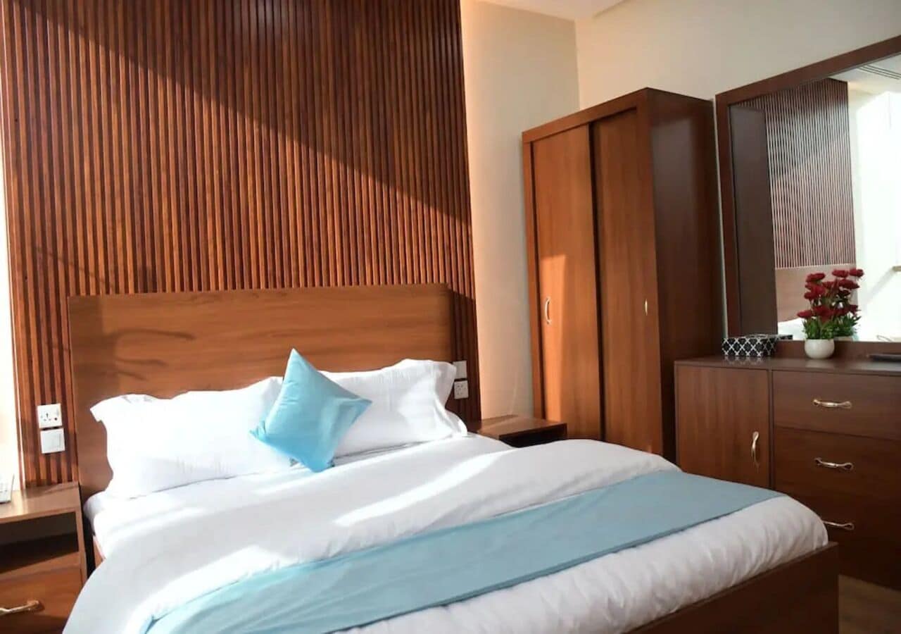 A hotel room with a wooden headboard, a bed with white bedding and a blue accent pillow, a wooden wardrobe, and a nightstand with a lamp.