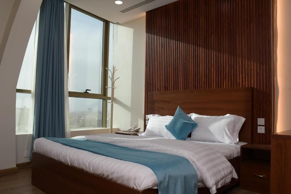 Une chambre d’hôtel moderne avec un grand lit, une tête de lit en bois, une table de chevet et une fenêtre avec des rideaux.