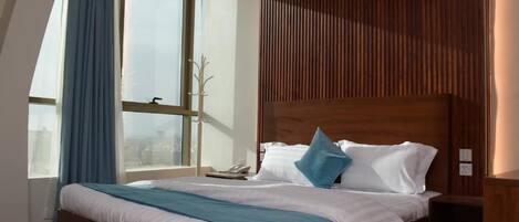 Ein modernes Hotelzimmer mit einem großen Bett, einem Holz-Kopfteil, einem Nachttisch und einem Fenster mit Vorhängen.
