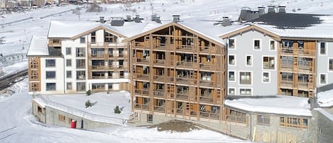 A multi-story hotel with a wooden facade, situated in a snowy mountainous area with ski slopes in the background.