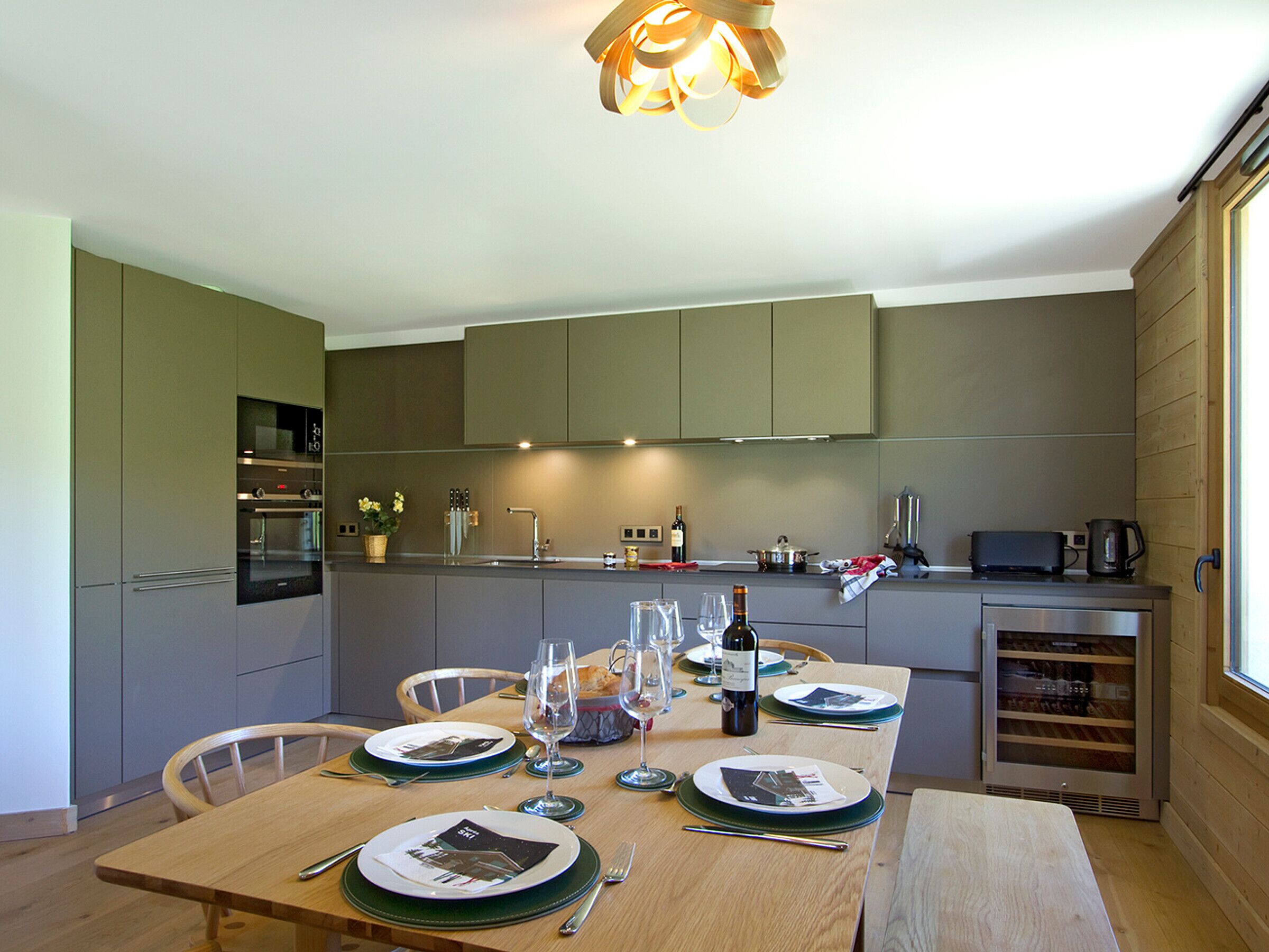 A modern kitchen with a dining area set for a meal, featuring a wooden table, chairs, and a built-in oven.