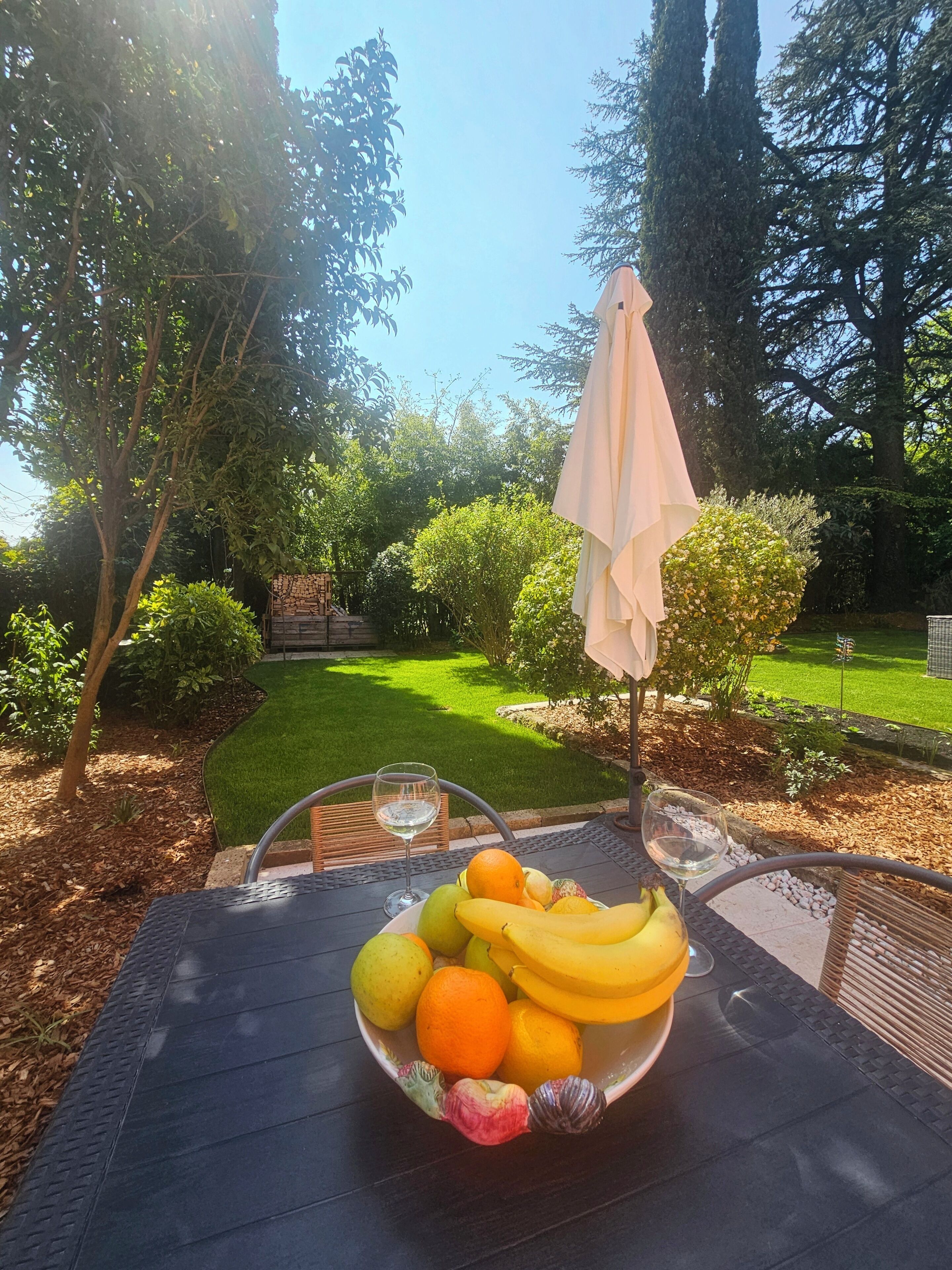 An outdoor table with a fruit bowl and two glasses of white wine, surrounded by a garden with a lawn and trees.