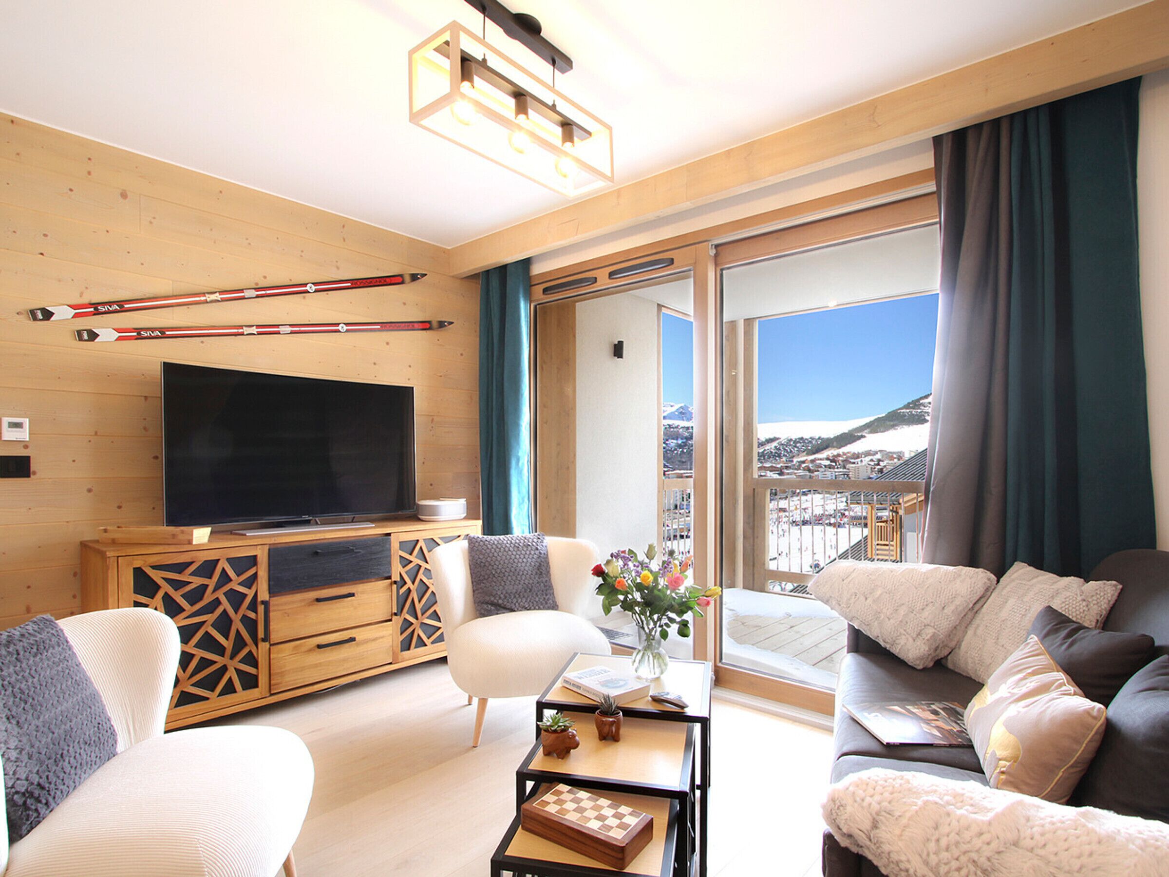 Ein modernes Wohnzimmer mit einem Flachbildfernseher, Holzmöbeln, einem Schachtisch und Blick auf eine verschneite Landschaft durch große Fenster.