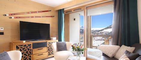 A modern living room with a flat-screen TV, wooden furniture, a chess table, and a view of a snowy landscape through large windows.