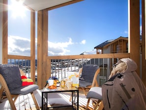 Una terraza de madera con dos sillas y una mesa, con vistas a un paisaje nevado con edificios y una montaña.