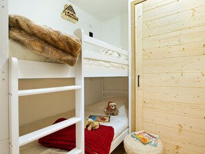 Uma cama beliche com um cobertor vermelho e um ursinho de pelúcia, uma pequena estante com uma casinha de brinquedo e um guarda-roupa de madeira.