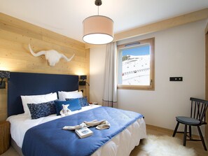 A bedroom with a wooden headboard, a large bed with blue bedding, a white stuffed animal, a book, and a window with a view of a snowy landscape. - Modern apartment at the foot of the slopes (Huez)