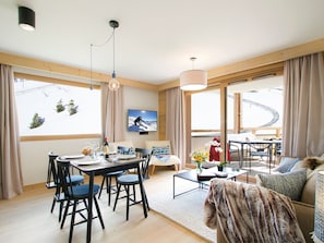 A modern living room with a dining area, large windows with curtains, a flat-screen TV, and a cozy seating area with a fur throw. - Modern apartment at the foot of the slopes (Huez)