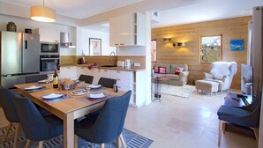 Eine moderne Küche mit Edelstahlgeräten, ein Essbereich mit Holztisch und Stühlen sowie ein Wohnbereich mit Sofa und Sesseln.