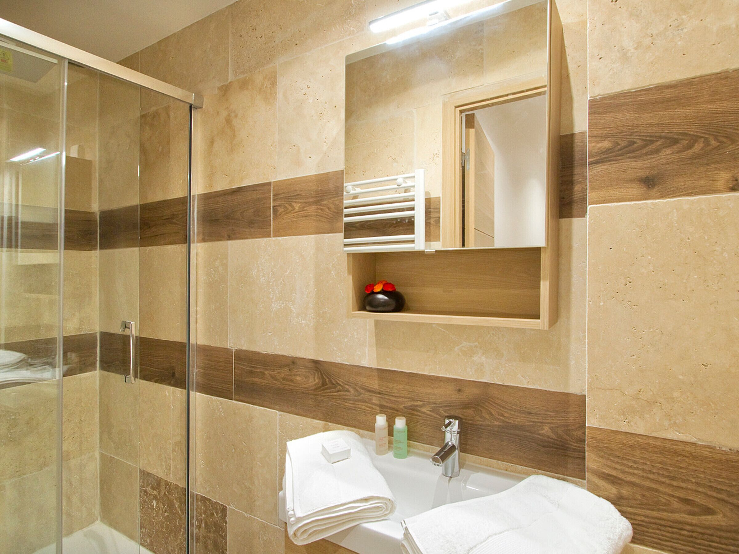 A modern bathroom with a glass shower enclosure, a built-in sink with a mirror, and a wooden shelf with toiletries.