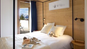 A bedroom with a bed, pillows, a stuffed toy, a bedside table with a book, and a view of mountains through an open door. - Spacious apartment with garden - 5 guests (Huez)