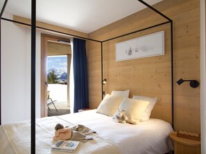 A bedroom with a bed, pillows, a stuffed toy, a bedside table with a book, and a view of mountains through an open door. - Spacious apartment with garden - 5 guests (Huez)