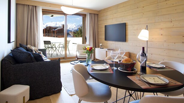 A modern living room with a sofa, dining table, chairs, and a TV mounted on the wall. - Spacious apartment with garden - 5 guests (Huez)