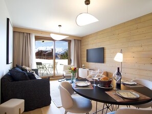 A modern living room with a sofa, dining table, chairs, and a TV mounted on the wall. - Spacious apartment with garden - 5 guests (Huez)
