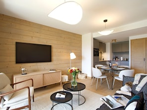 A modern living room with a flat-screen TV, a dining area with a table and chairs, and a kitchen in the background. - Spacious apartment with garden - 5 guests (Huez)