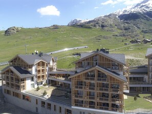A modern hotel with multiple floors and large windows, set against a backdrop of green hills and a mountain range. - Spacious apartment with garden - 5 guests (Huez)