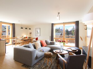 Ein modernes Wohnzimmer mit einer grauen Couch, einem Essbereich mit Holzmöbeln und großen Fenstern mit Blick auf die Berge.