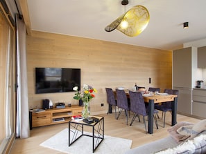 Una sala de estar moderna con piso de madera, una mesa de comedor con sillas, un televisor de pantalla plana montado en la pared y una lámpara decorativa.