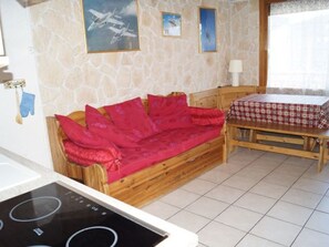 A room with a stone wall, a wooden sofa with red cushions, a wooden table with a checkered tablecloth, and a window with curtains. - Duplex with parking - 4 guests, stunning view (Les Deux Alpes)